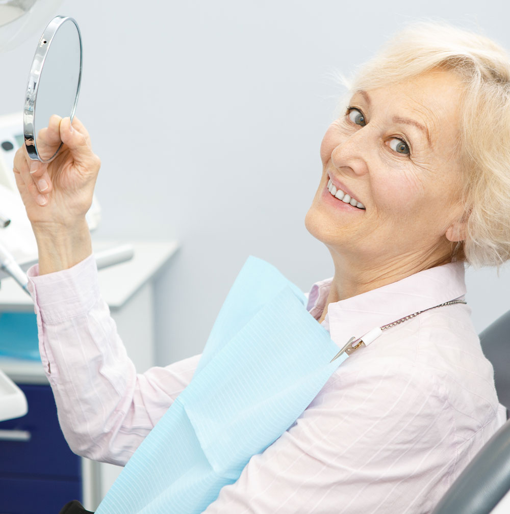 senior woman holding up a mirror to view her tooth replacement at the dentist office
